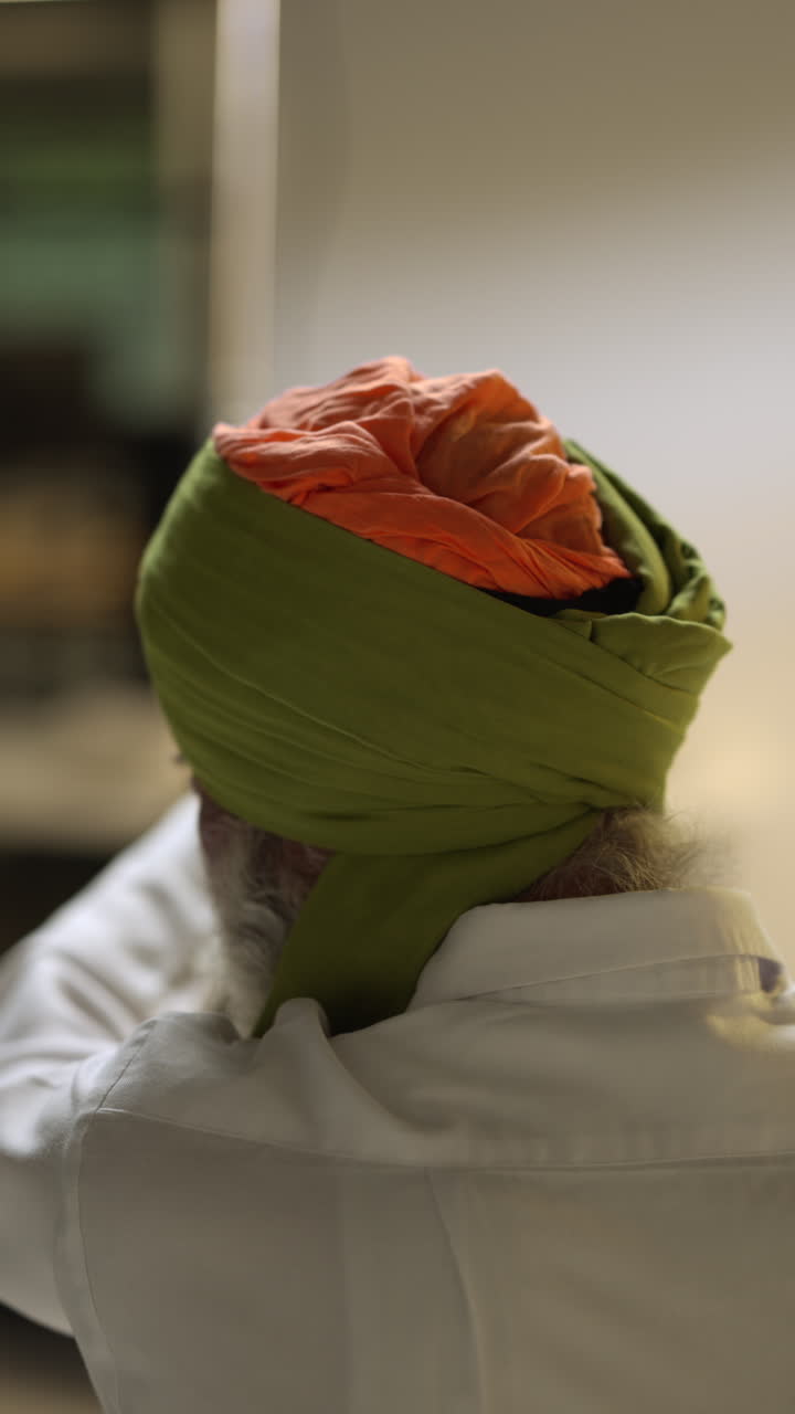 una toma de video vertical de un anciano sikh con barba atando tela para un turbante mirándose en el espejo desde atrás
