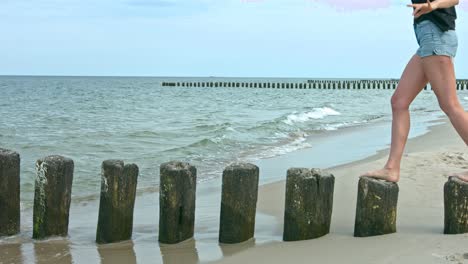 A-woman-standing-on-top-of-a-wooden-post,-walking-from-one-to-another-at-the-beach-as-the-waves-roll-in