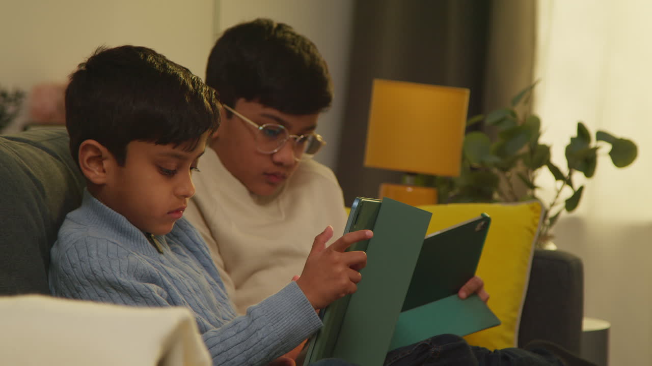 Two Young Boys Sitting On Sofa At Home Playing Games Or Streaming Onto Digital Tablets