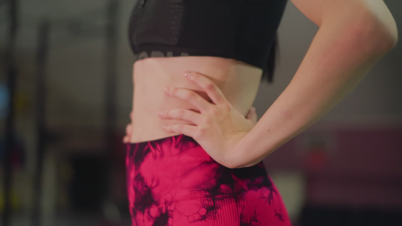 closeup female torso in gym wearing pink leggings and black sports bra, hand resting on hip with tiedye print, emphasis on core strength and posture, studio lights and window bokeh, confident pose