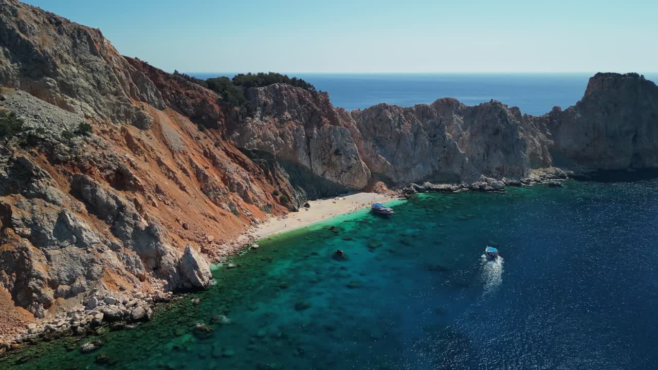 Vista aérea con dron de una playa escondida en la isla de Suluada en Turquía, rodeada de escarpados acantilados rocosos y aguas mediterráneas claras de color turquesa con un pequeño barco acercándose a la bahía