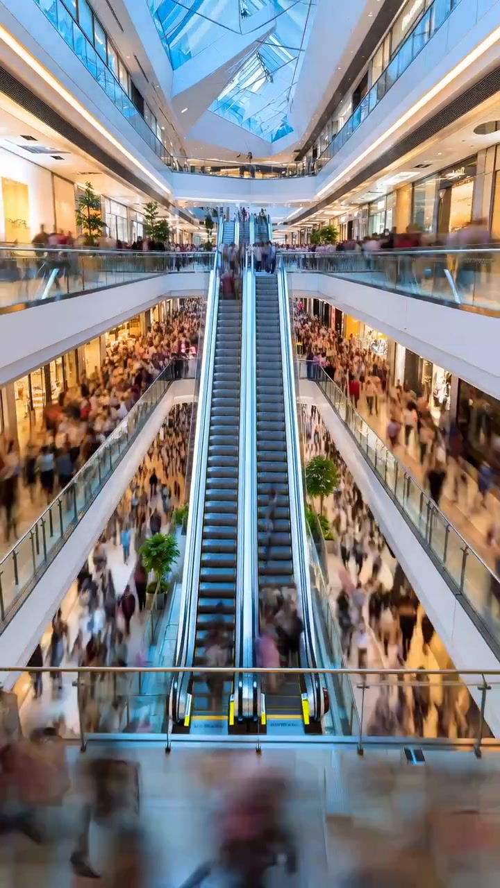 Højt vinkelperspektiv af et moderne indkøbscenterinteriør med centrale tvillingrulletrapper, slørede menneskemængder, mezzanin-niveauer, ovenlys, glasbalustrader, polerede refleksioner og en livlig detailhandelsatmosfære.