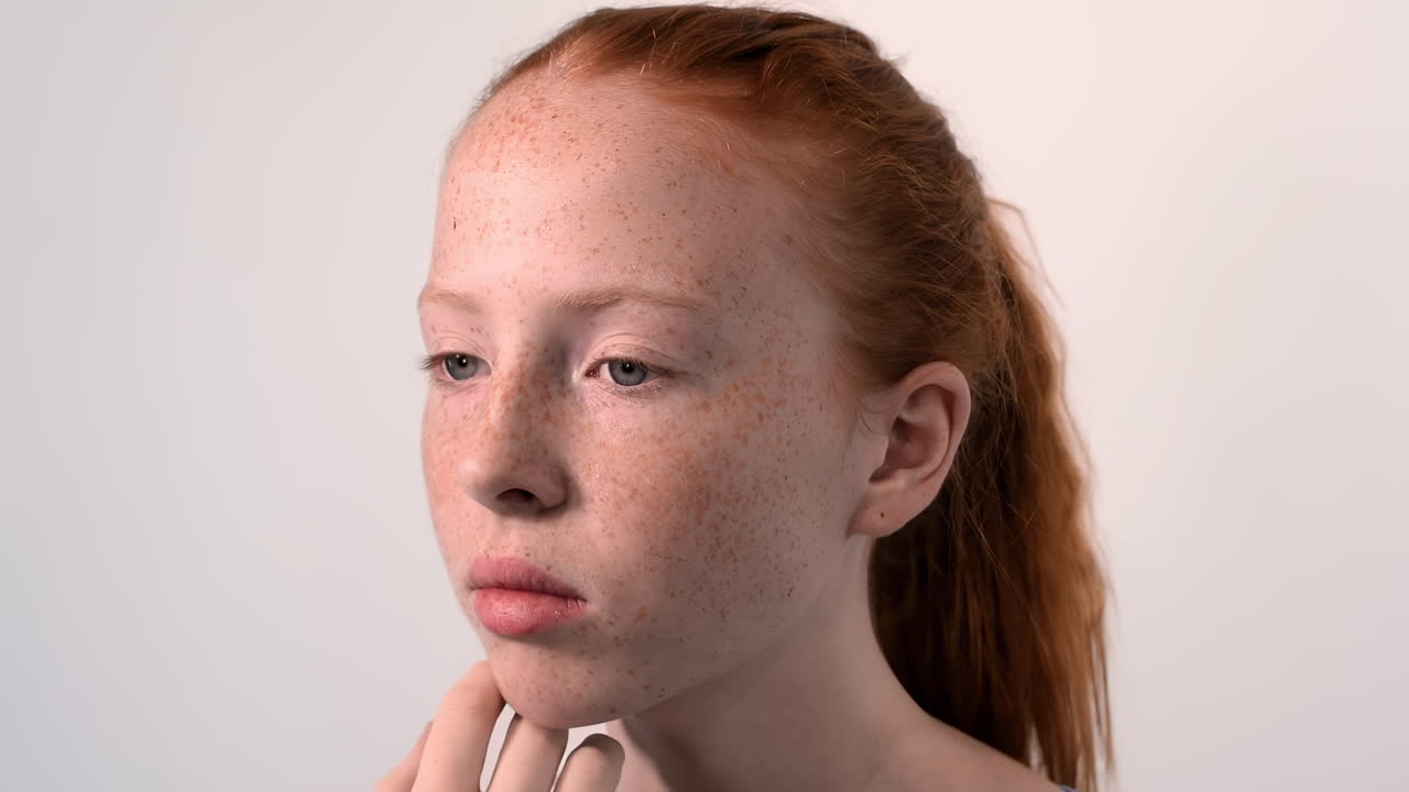 Red haired girl with blue eyes touching face full of freckles. Looking ahead. White background. Tighten hairs