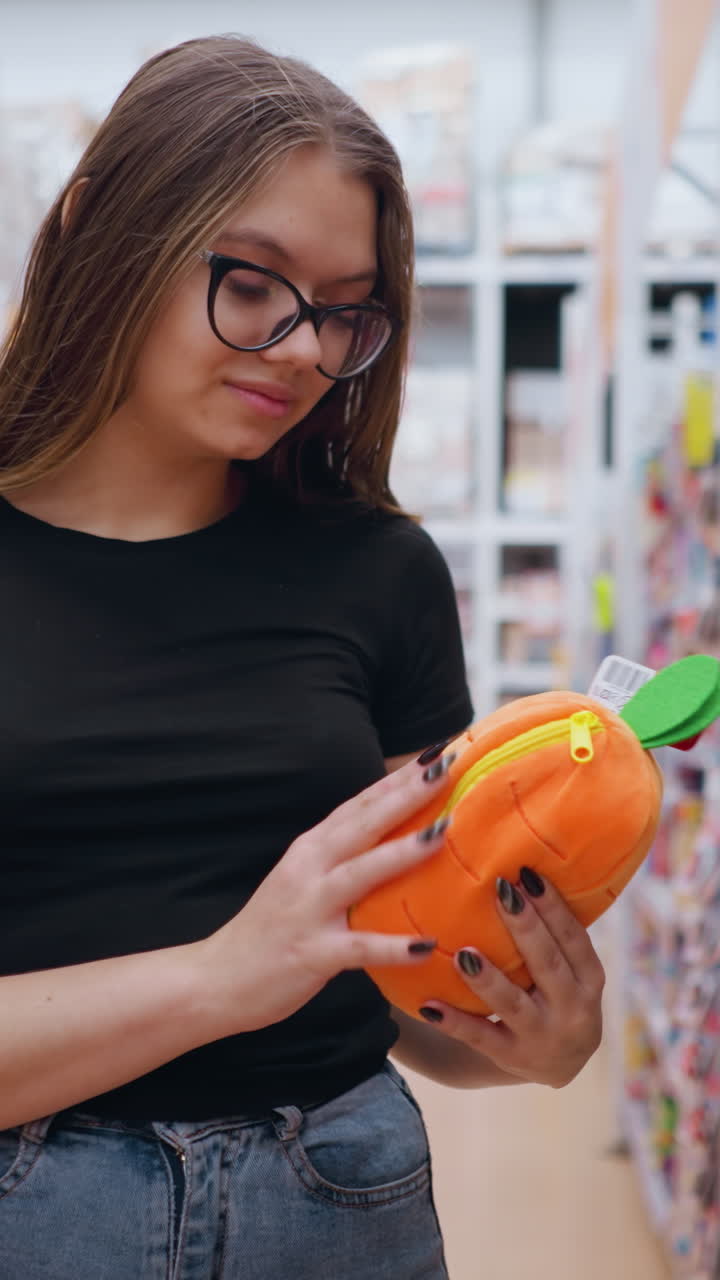 검은 티셔츠와 청바지를 입은 젊은 여성이 잘 밝은 쇼핑몰의 상점 선반에서 밝은 오렌지색 플러시 가방을 집어 들고, 그녀는 다채로운 액세서리로 가득 찬 장난감 섹션을 탐색하는 동안 항목을 조사합니다.