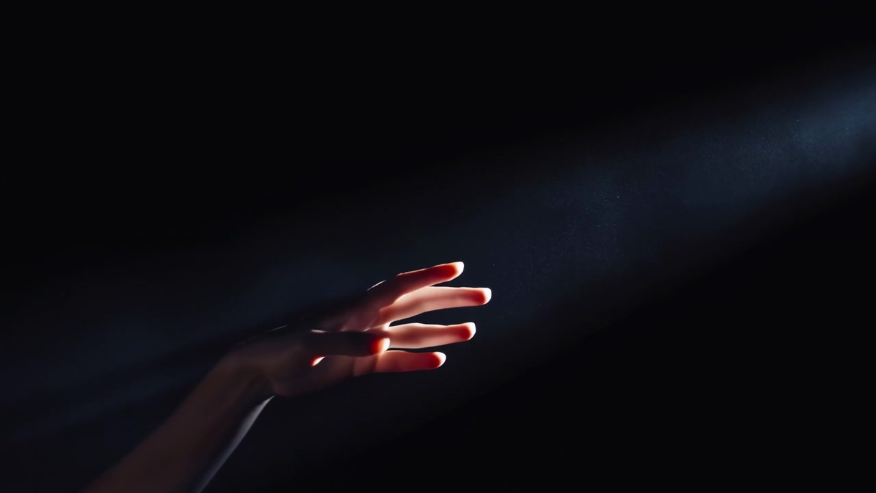 Left hand and forearm move into cool diagonal light from lower left, palm up and fingers spread as fingertips glow with rim highlights against dark background, creating moody contrast