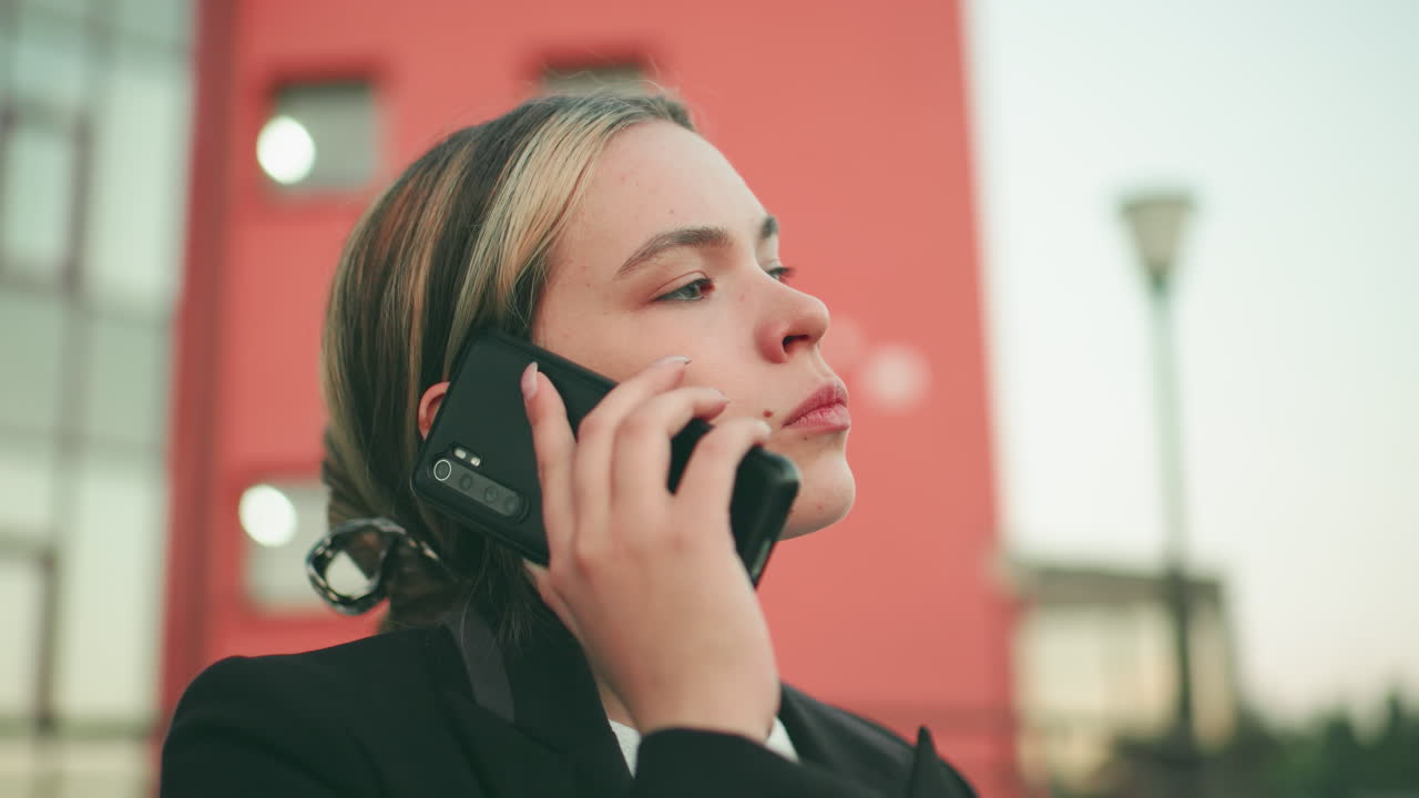 Lady stressed during phone call in black suit wiping forehead, showing frustration and exhaustion, with modern building slightly blurred in background
