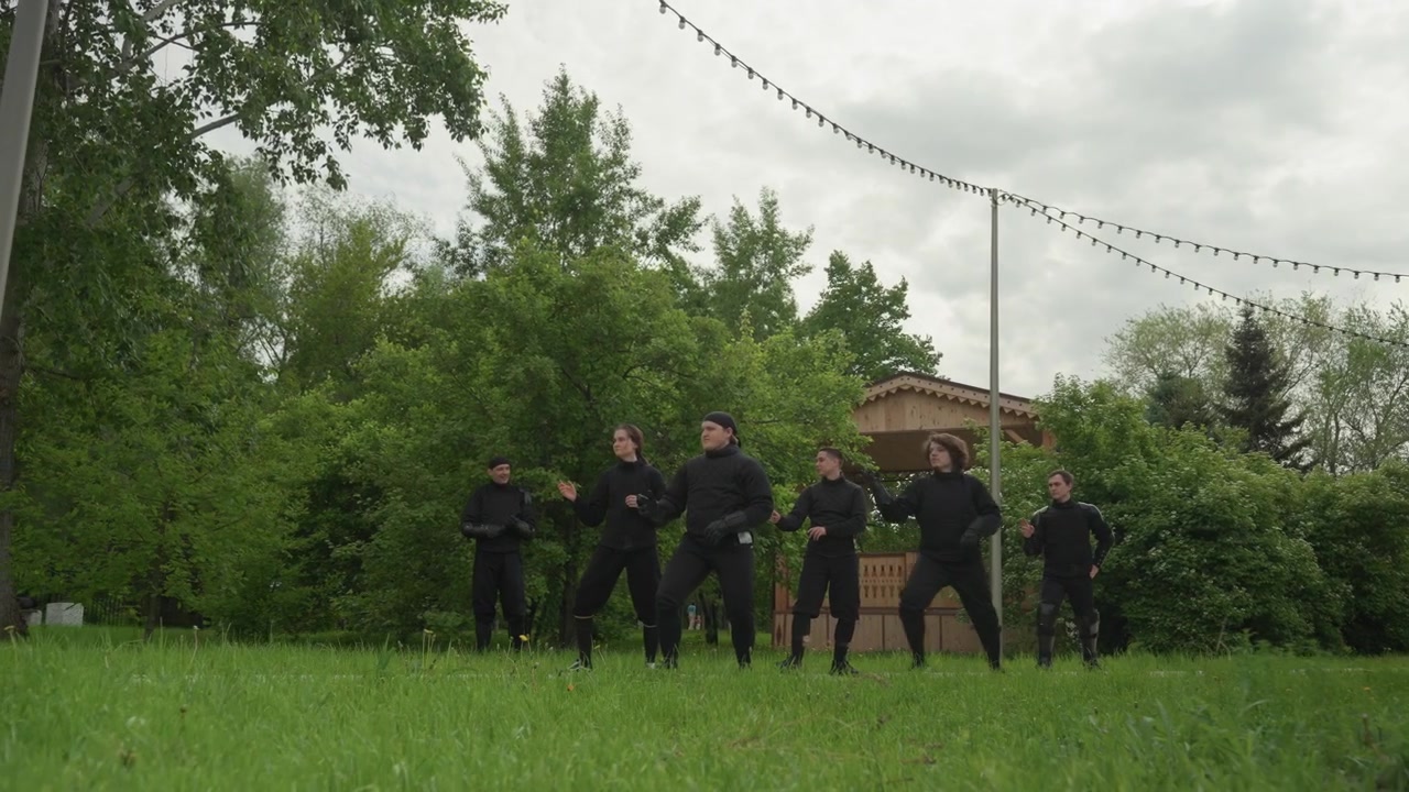 Gruppo di persone in abiti neri che eseguono movimenti sincronizzati all'aperto sull'erba, con alberi e una struttura in legno sullo sfondo. Le persone mantengono gesti coordinati per tutto il tempo, suggerendo.
