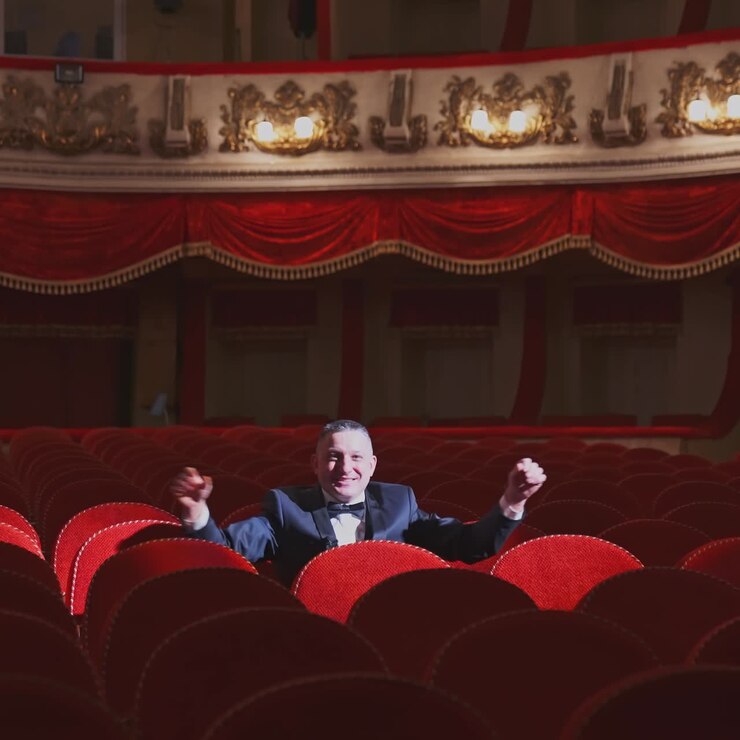 Successful man in suit alone in large auditorium. Actor man is sitting among empty red armchairs and singing in opera house.
