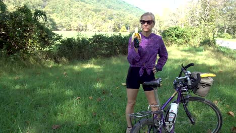 mujer bonita, rubia y madura con gafas de sol y guantes al lado de su bicicleta comiendo un plátano al lado de la carretera