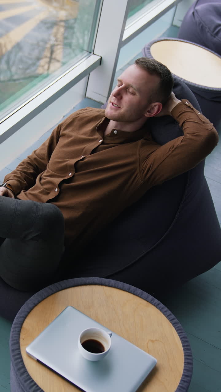 Businessman relaxing in office. Young man sitting in comfortable armchair with closed eyes and dreaming at daytime next to the window with city view. Top view. Vertical video