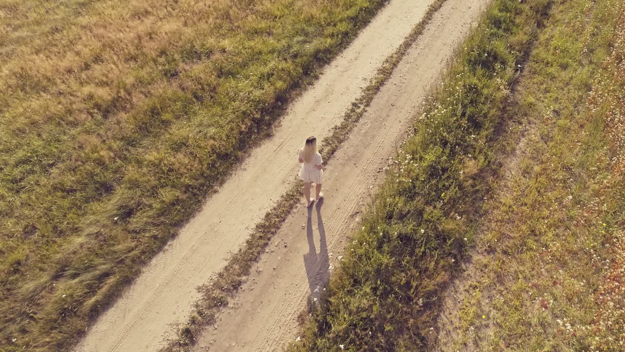 An aerial tracking shot of a woman wearing a white dress and holding a flower walking down a dirt track