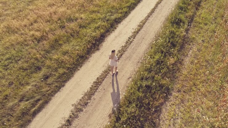 An aerial tracking shot of a woman wearing a white dress and holding a flower walking down a dirt track