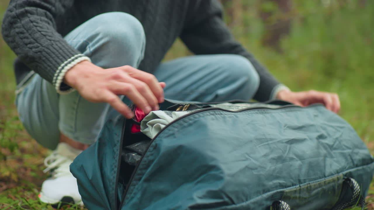 close up of camper squatting on grassy forest floor opening green tent bag and setting it down beside him, preparing for outdoor activity in woodland setting with visible hands and sneakers