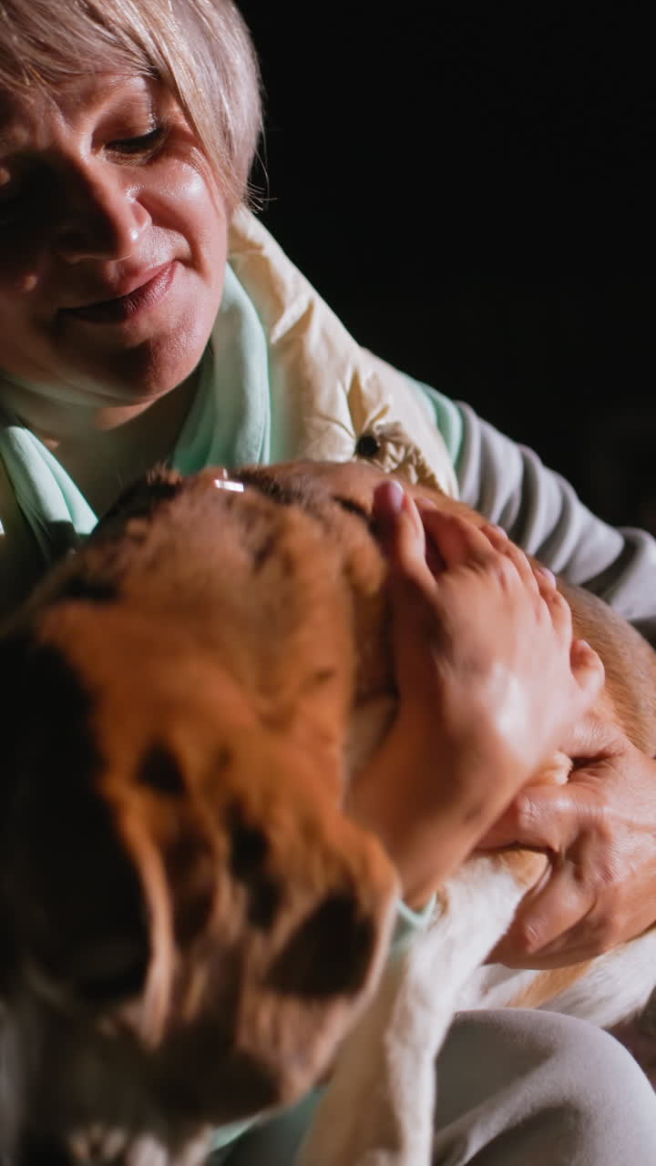 Mujer blanca abrazando a un beagle por la noche; retrato de primer plano que muestra una caricia suave con la mano, sudadera color menta, chaleco color crema, mirada cálida, iluminación discreta, consuelo emocional, dueña de mascota jubilada, vínculo íntimo