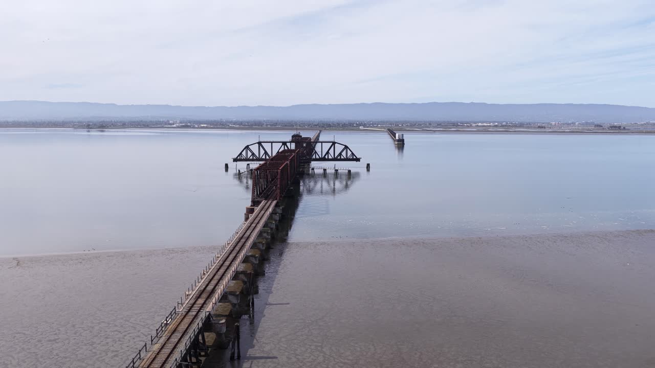 Drone footage flying over the historic and abandoned Dumbarton Rail Bridge, showing the old truss and swing bridge structure reflecting in the calm waters of the San Francisco Bay, California