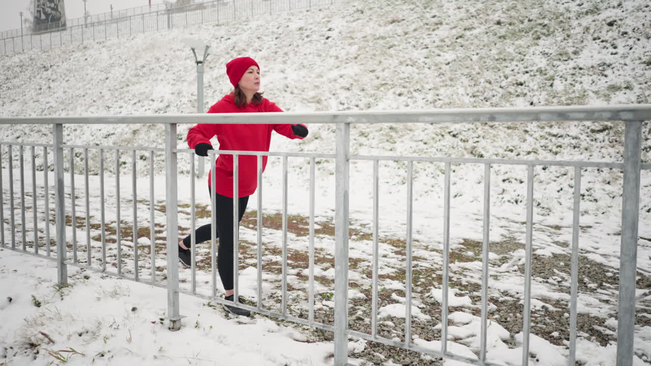 겨울 동안 야외에서 운동하는 여성, 다리를 밖으로 으면서 철의 레일링을 들고, 빨간 옷을 입고, 배경은 눈 인 땅, 안개 분위기, 멀리 있는 나무입니다.