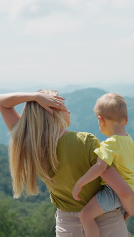 woman holds little son and adjusts long loose hair looking at distant mountains silhouettes at eco resort backside view slow motion