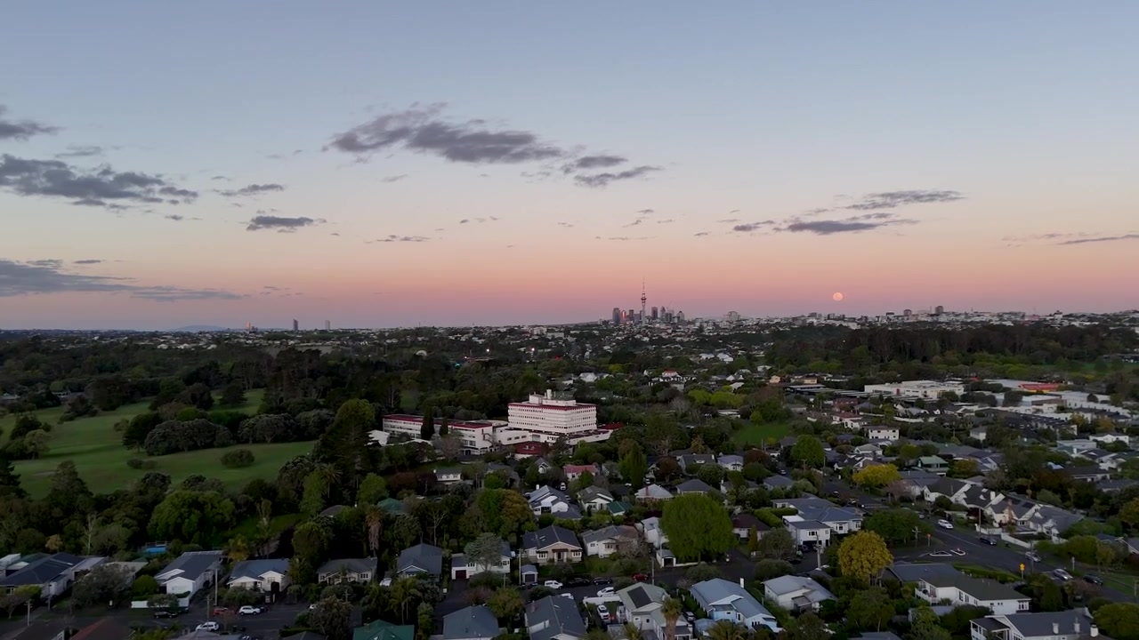 Full moon rises over faint Auckland skyline at dusk, suburban rooftops and tall white apartment building spread across tree filled foreground neighborhood in New Zealand. Aerial drone rising wide shot