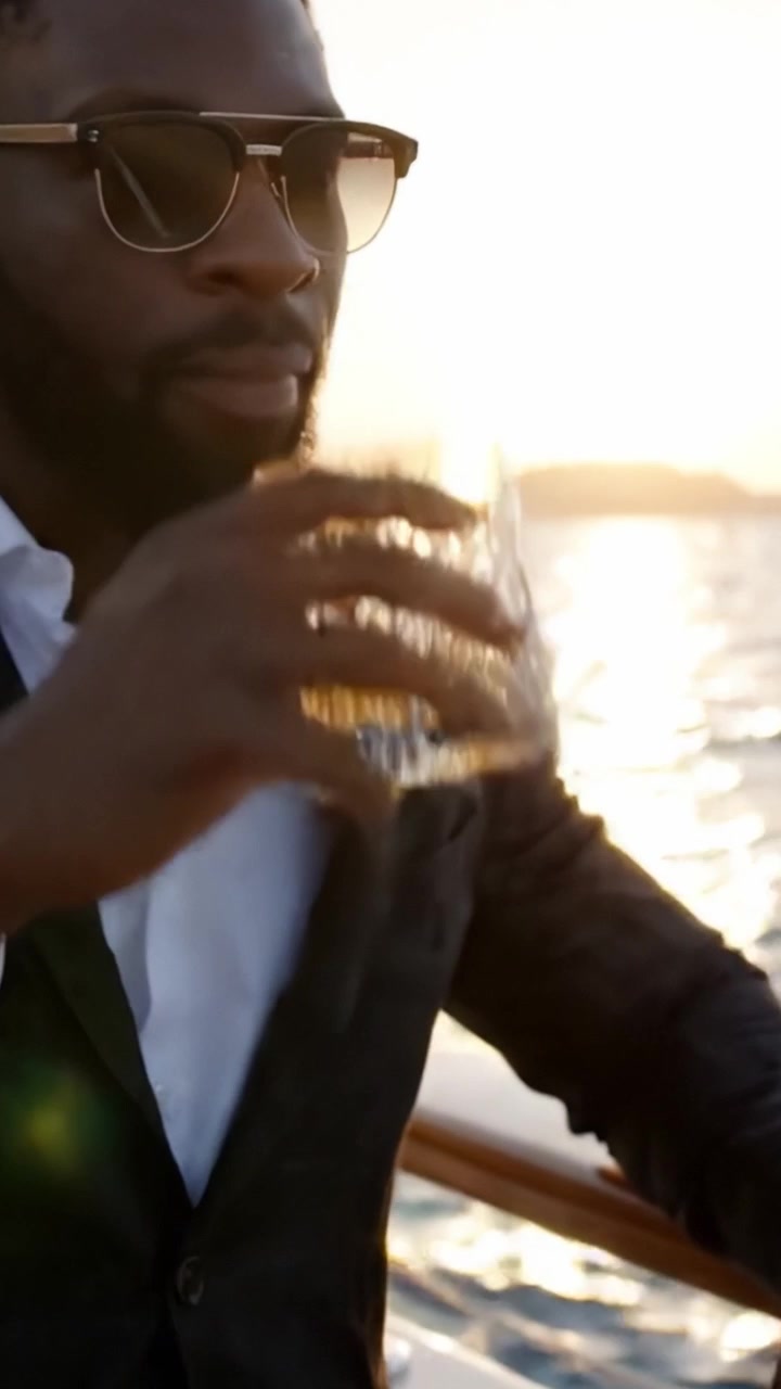 Un uomo elegante si gode una serata raffinata su uno yacht con un bicchiere di whisky in mano, ammirando un tramonto mozzafiato sull'acqua.