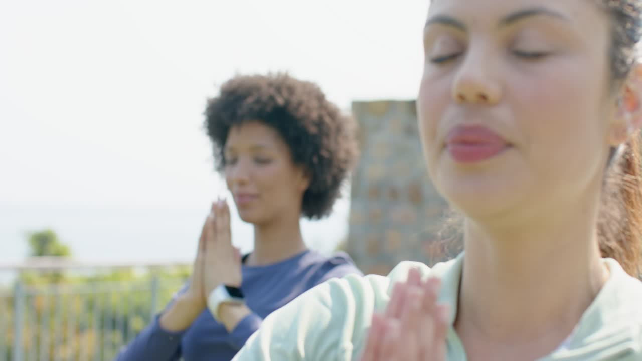 Diverse amiche con smartwatch che sollevano le braccia in posizione di preghiera per la meditazione sul balcone.
