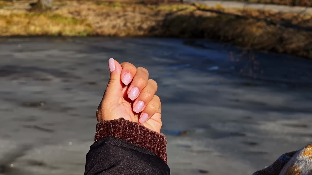 Primo piano di una mano che mostra unghie acriliche rosa lucide su uno sfondo di acqua ghiacciata e un ambiente esterno soleggiato