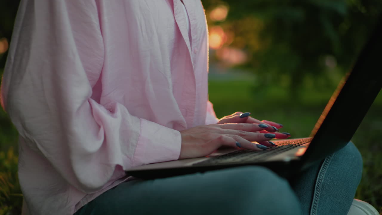 primer plano de una mujer con camisa rosa escribiendo en una computadora portátil con uñas bien pulidas, iluminadas por la luz solar suave, el fondo presenta vegetación con un hermoso efecto de luz bokeh