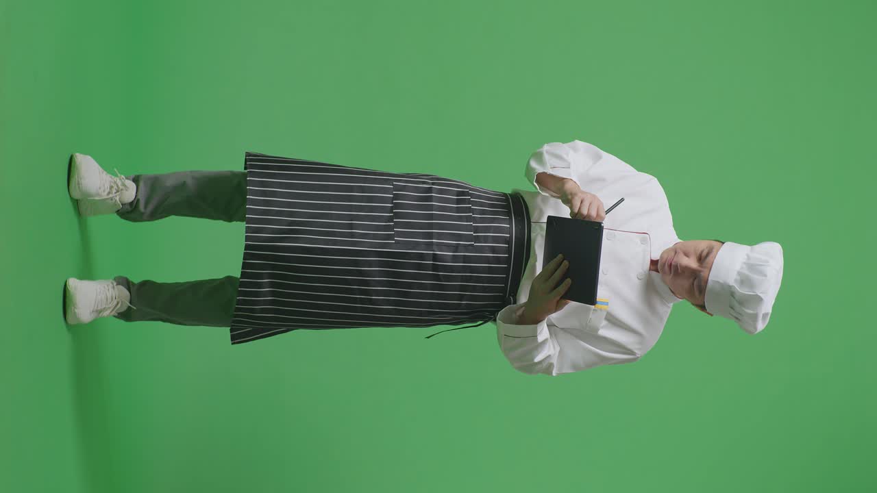 cuerpo lleno de cocinero hombre asiático tomando nota en una tableta mientras estaba de pie en el estudio de fondo de la pantalla verde