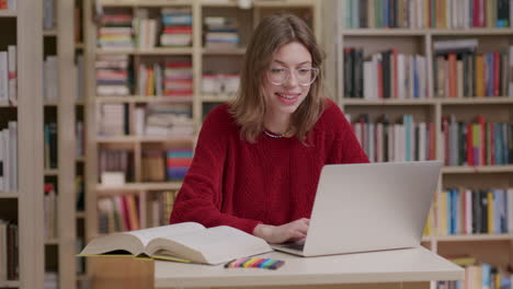 una dama caucásica sonriente con suéter rojo y gafas trabaja en una laptop en la biblioteca
