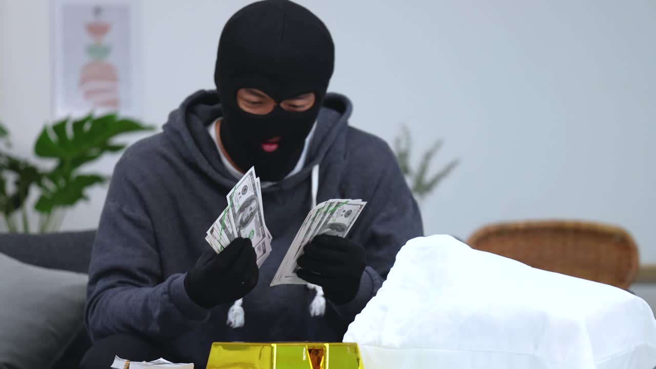 A masked thief wearing black gloves counts stacks of dollar bills after a burglary