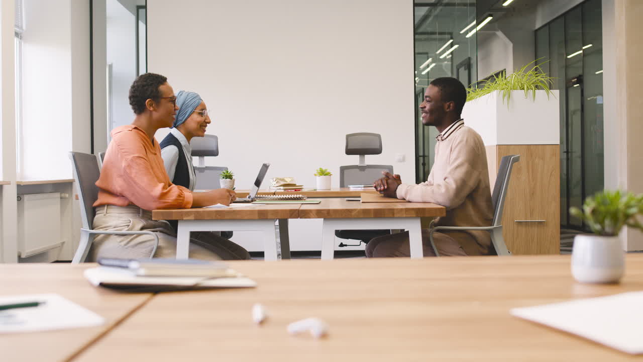 An Woman And A Muslim Woman Co Workers Interview A Young Man Sitting At A Table In The Office 11