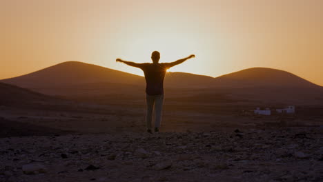 happy person raising arms and walking towards sunset at goldenhour with mountain range in the back