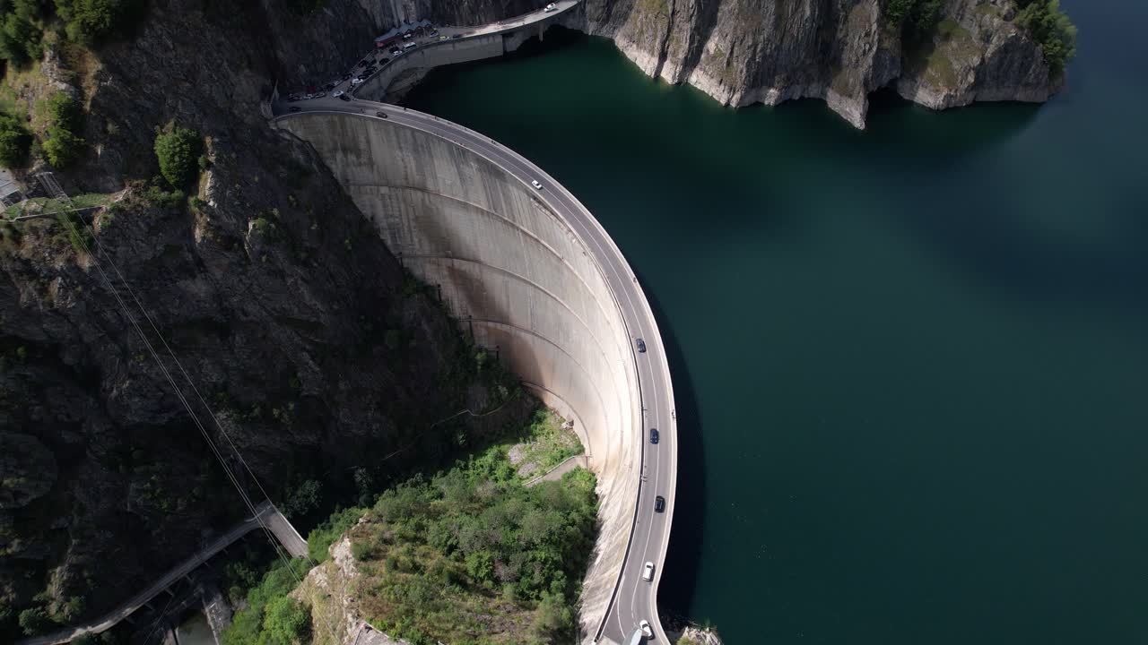 Drone shot of cars driving on Vidraru Dam in Romania, circling left. Blue lake, dramatic curves, and green mountain backdrop in 4K.