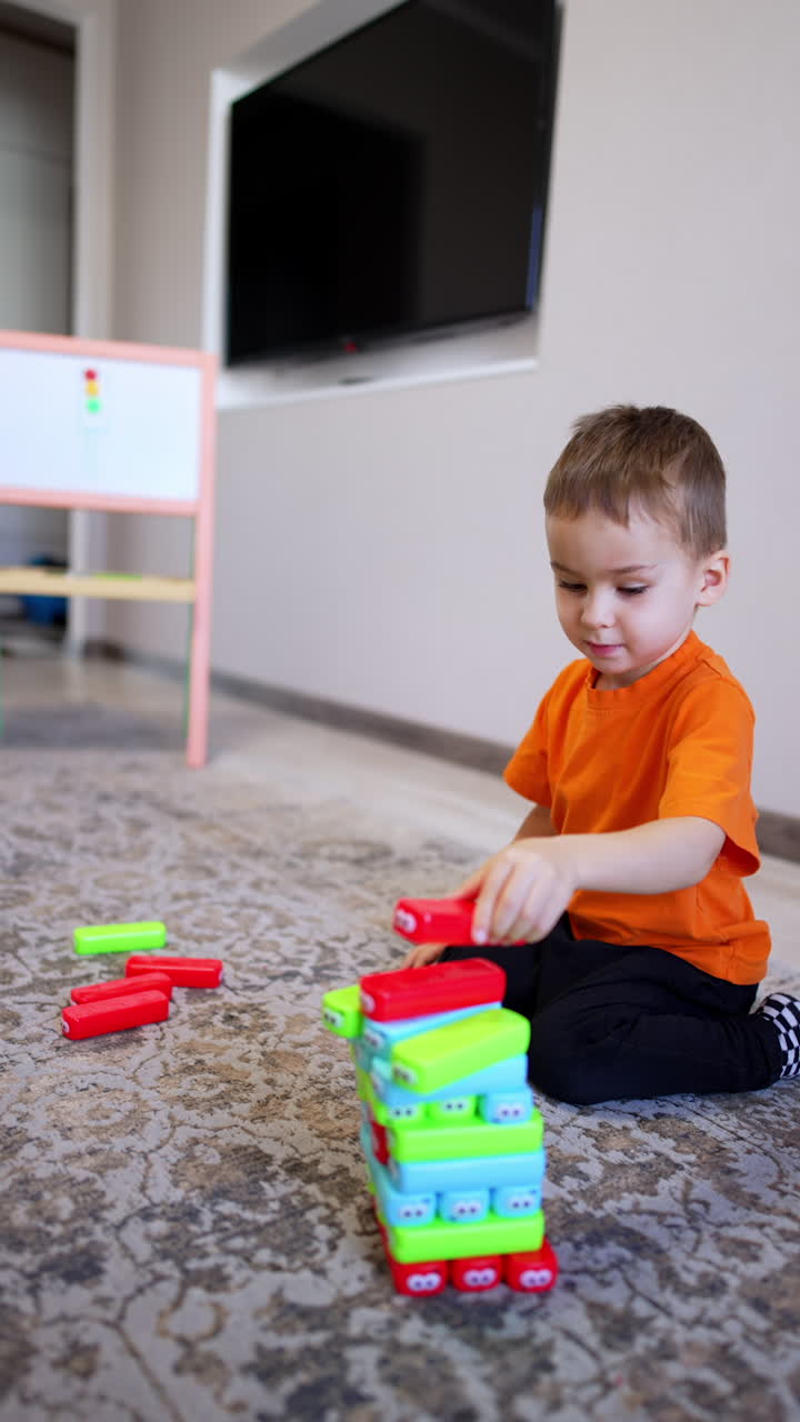 Four-year-old toddler in orange t-shirt plays with toys on the floor. Kid builds a tower of colorful blocks. Vertical video.