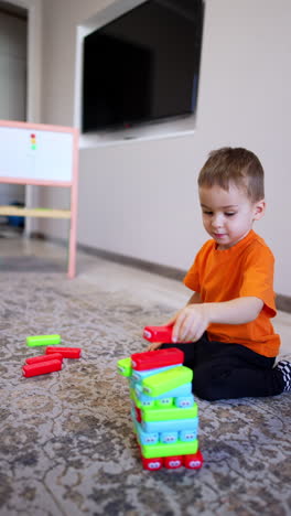 four-year-old toddler in orange t-shirt plays with toys on the floor. kid builds a tower of colorful blocks. vertical video.