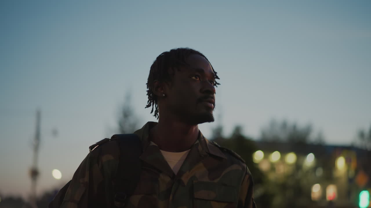 Black ExSoldier Walking Through City Dusk, Camouflage Jacket And Backpack, Blurred Lights And Skyline Behind, Steady Forward Gaze, Cinematic Night Bokeh, Hopeful Return To Civilian Life