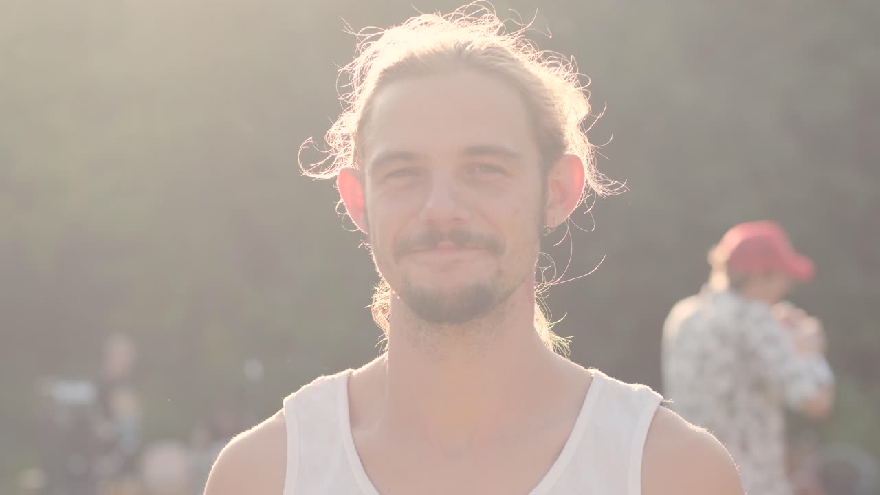 해가 지는 배경에 카메라를 향해 미소 짓는 긴 머리카락을 가진 젊은 잘생긴 스터 남자의 초상화