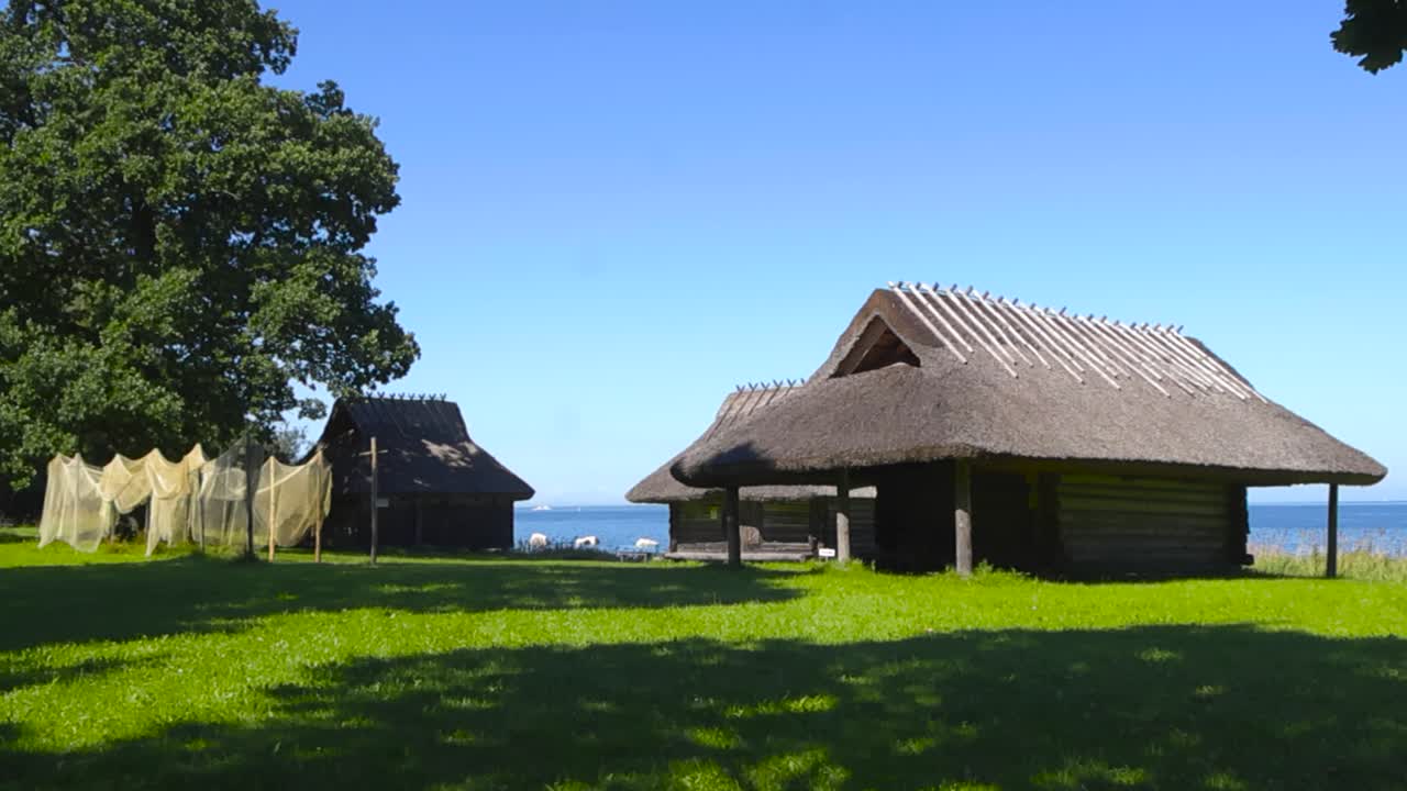 Rural seaside shoreline traditional and retro fishing village with fishing nets hanging on wooden poles and log cabins with thatched reed and straw roofs are on the sunny and grassy beach, horizon