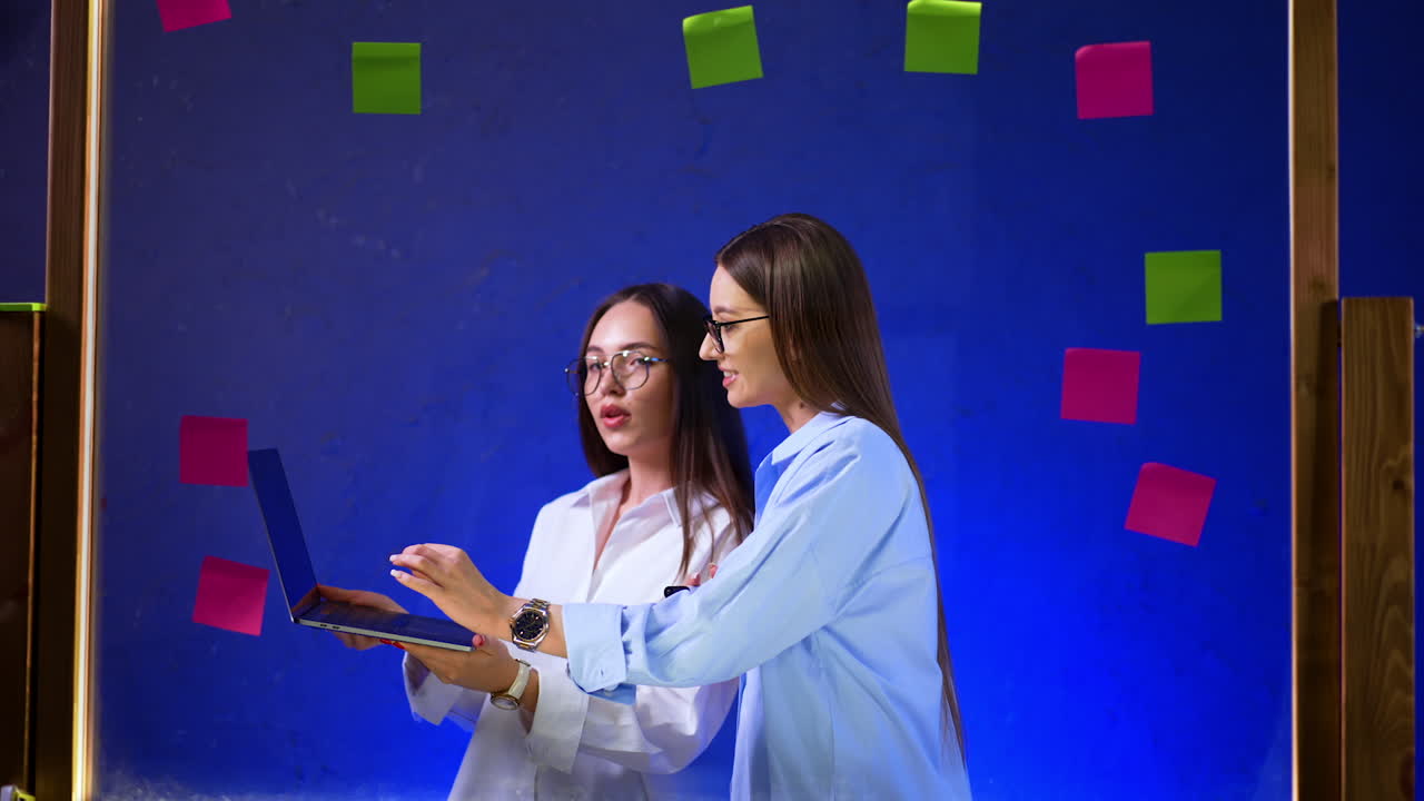 Women working together on laptops. Two women discuss ideas while working together with a laptop, surrounded by colorful sticky notes on a blue background