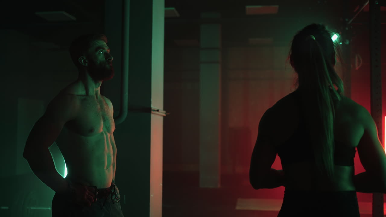 Spor salonunda iki adam ve bir kadın, karanlık neon ışığında yatay barda birlikte barfiks çekiyor. Fitness ve antrenman atmosferi.