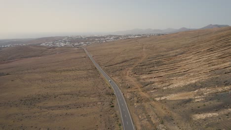 Drone-flight-over-the-road-leading-through-volcanic-island-landscape,-Lanzarote,-Canary-Island