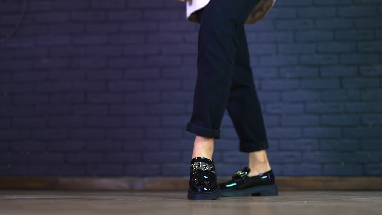 Girl in black jeans demonstrating modern massive shoes with silver chain decoration. Grey brick wall background.