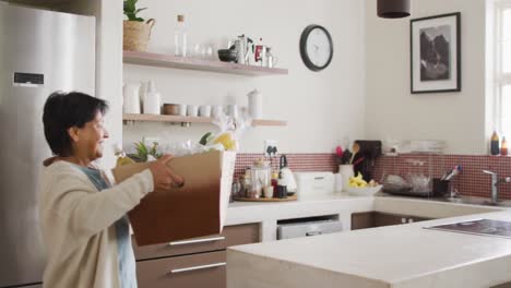 Smiling-senior-biracial-woman-holding-box-of-vegetables-in-kitchen-alone