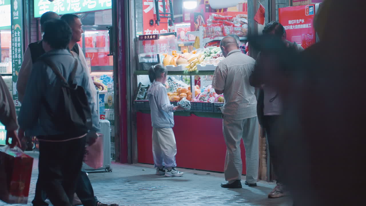 夜の路上にあるコンビニエンスストアで、アジア系の店主と子供がネオンの棚を眺めている。地元の買い物客や観光客が列をなし、濡れた舗道にはシアンとピンクの光が反射している。飾らない都市の交流が垣間見える。