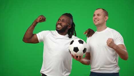 excited african american and caucasian teammates rejoicing victory standing with ball at chromakey background. portrait of satisfied happy young sportsmen gesturing looking away on green screen.