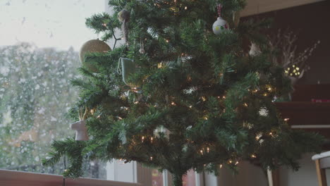 a christmas tree covered in lights and decorations next to a window through snow can be seen falling on the other side