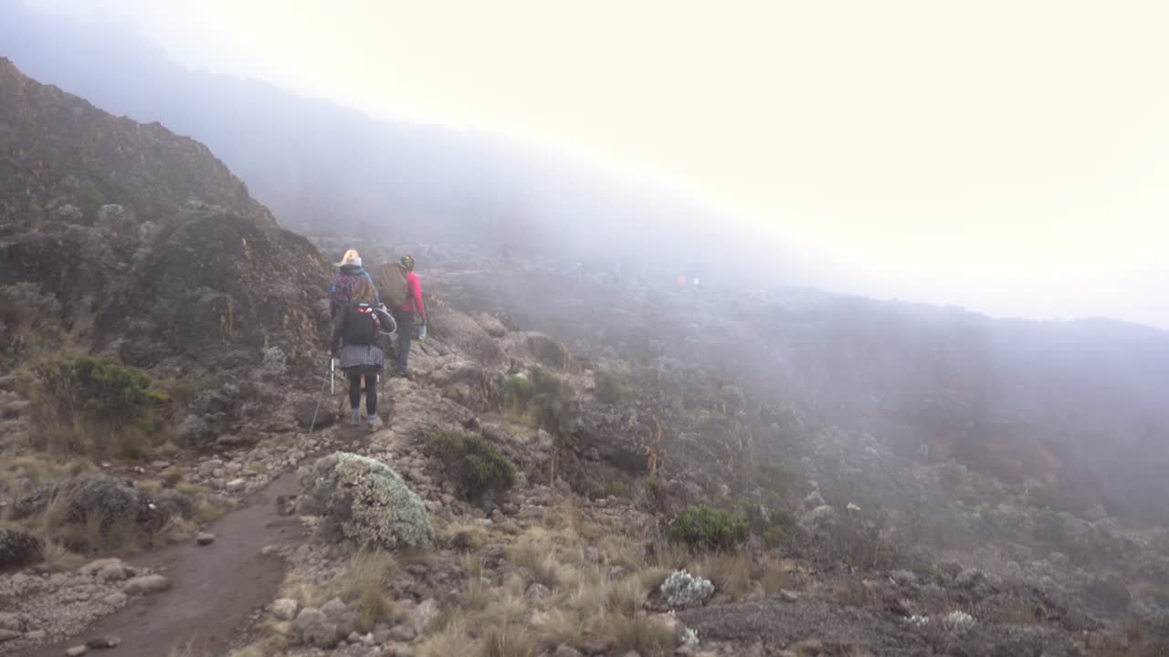 toma panorámica de dos excursionistas y un guía en el monte kilimanjaro caminando a través de nubes brumosas con otros excursionistas en backgorund