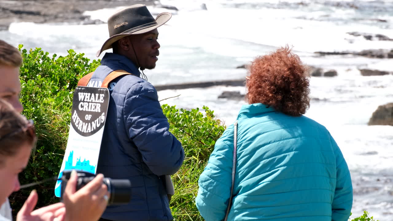 Female tourist chats with Hermanus Whale Crier on coastline, whale watching
