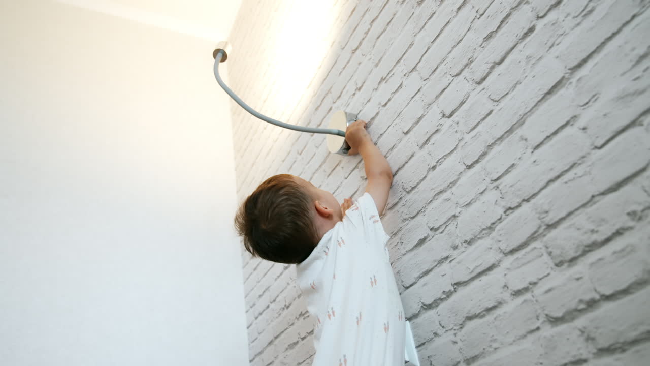 Little cute boy stands holding by the white brick wall. Curious toddler reaches the lamp above him to switch it off and on again. Low angle view.