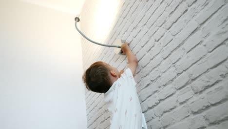 little cute boy stands holding by the white brick wall. curious toddler reaches the lamp above him to switch it off and on again. low angle view.