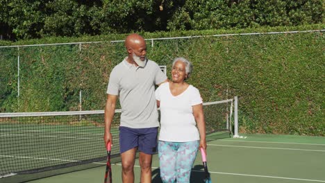 african american senior couple holding rackets walking on the tennis court